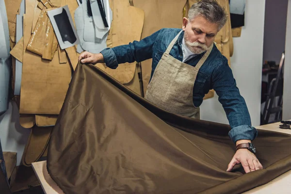 スタジオで作業テーブルに布を置く中間の老化させた男性ハンドバッグ職人 — ストック写真