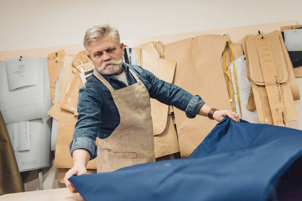 серьезный мужчина среднего возраста ремесленник сумки положить ткань на рабочий стол в студии
