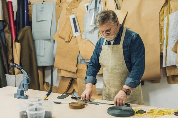 middle aged male handbag craftsman in apron and eyeglasses working with fabric at studio