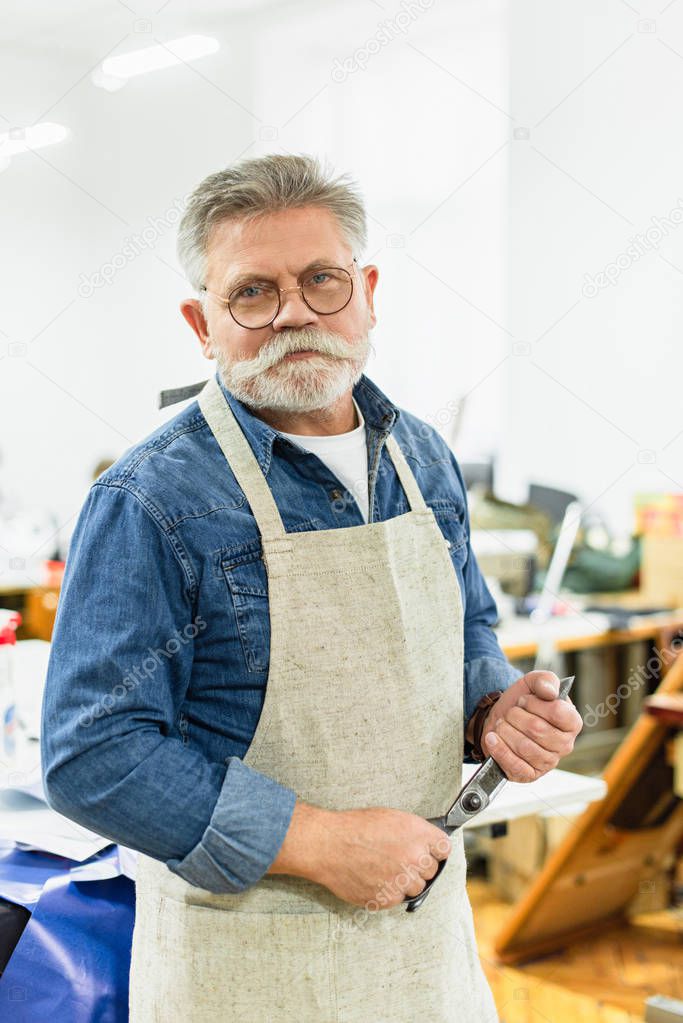 smiling middle aged male craftsman in apron posing with scissors at workshop