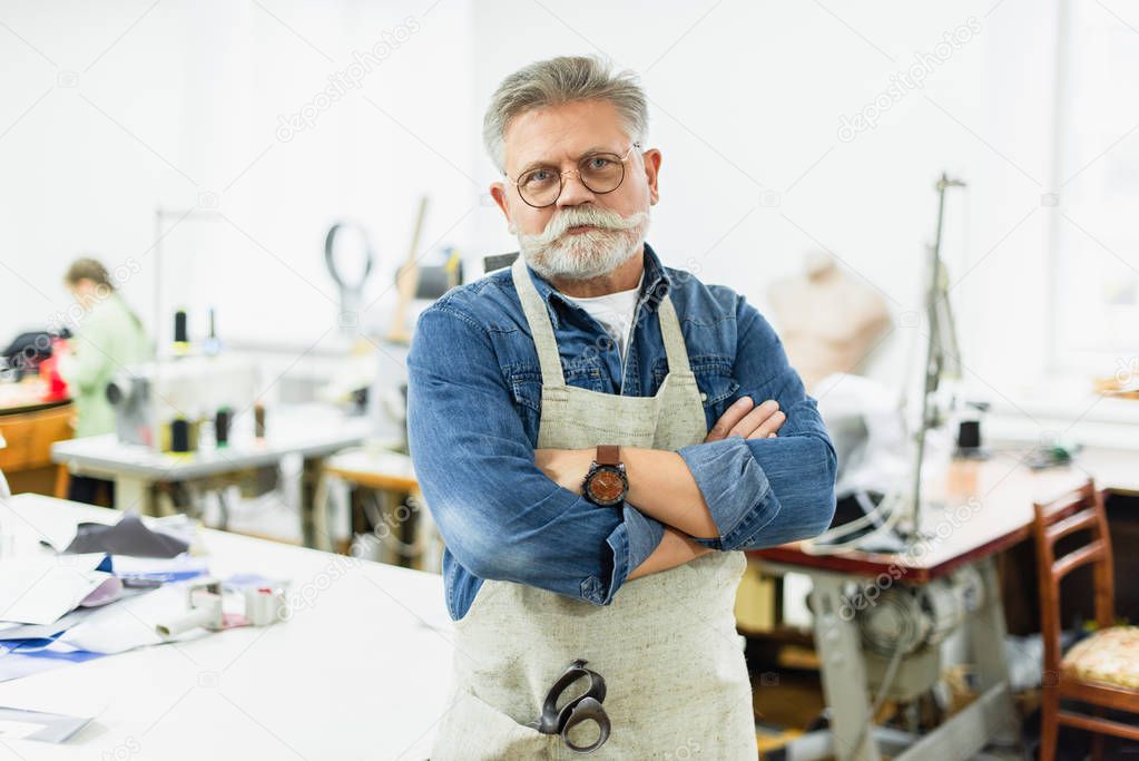 serious mature male craftsman in apron posing with crossed arms at workshop