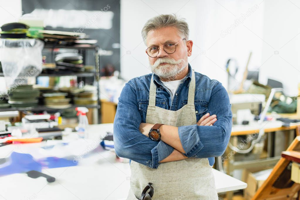 mature male craftsman in apron posing with crossed arms at workshop