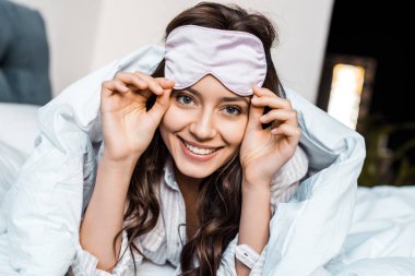 attractive smiling girl in sleeping eye mask lying in bed clipart