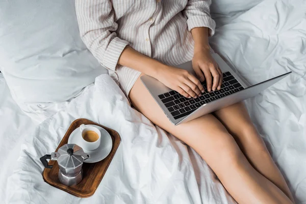 朝のベッドでノート パソコンを使ってコーヒーを持つ女性のトリミング ビュー — ストック写真