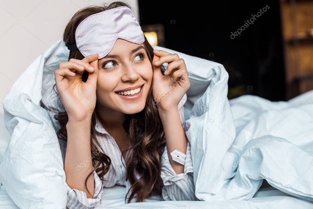cheerful girl in sleeping eye mask lying under blanket in bed