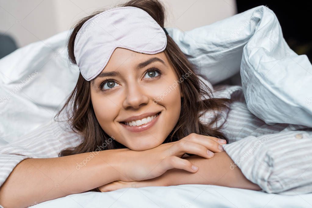 smiling dreamy girl in sleeping mask resting under blanket on bed