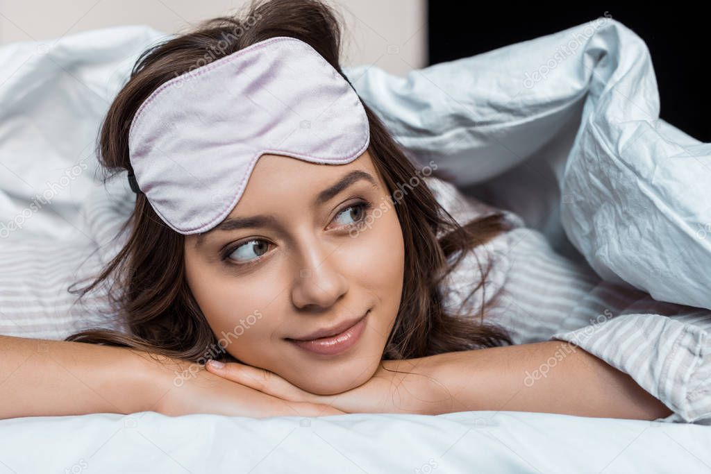 attractive girl in sleeping mask relaxing under blanket on bed  