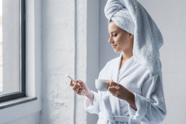 attractive young woman in bathrobe holding cup of coffee and using smartphone near the window clipart