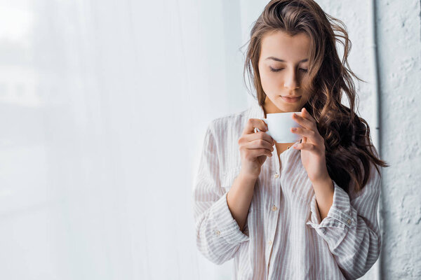 young brunette woman standing with cup of coffee near window in the morning 