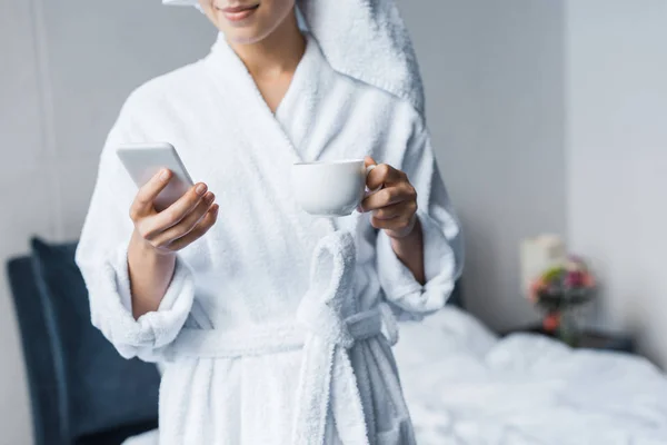 バスローブ一杯のコーヒーを保持している 朝の寝室でスマート フォンを使用して女性のトリミング ビュー  — 無料ストックフォト