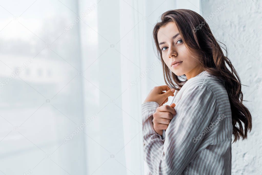 attractive tender girl in pajamas standing near window with white curtain 
