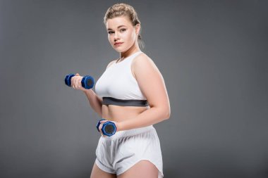 Genç boyutu artı dumbbells ile egzersiz ve gri izole kamera bakarak kadın