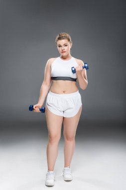 büyük boy kadında genç dumbbells ile egzersiz ve kameraya gri seyir spor giyim 