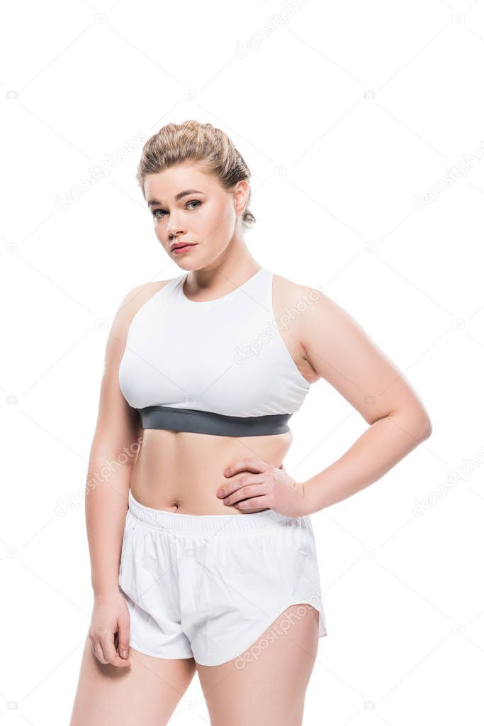 young overweight woman in sportswear standing with hand on waist and looking at camera isolated on white