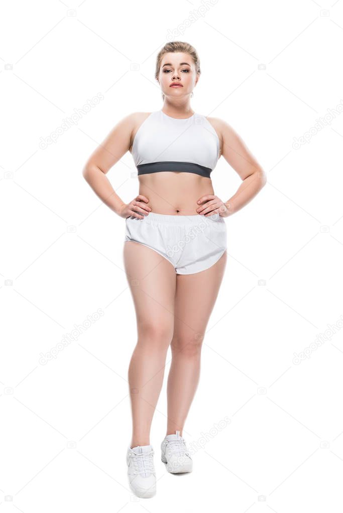 full length view of young size plus woman in sportswear standing with hands on waist and looking at camera isolated on white