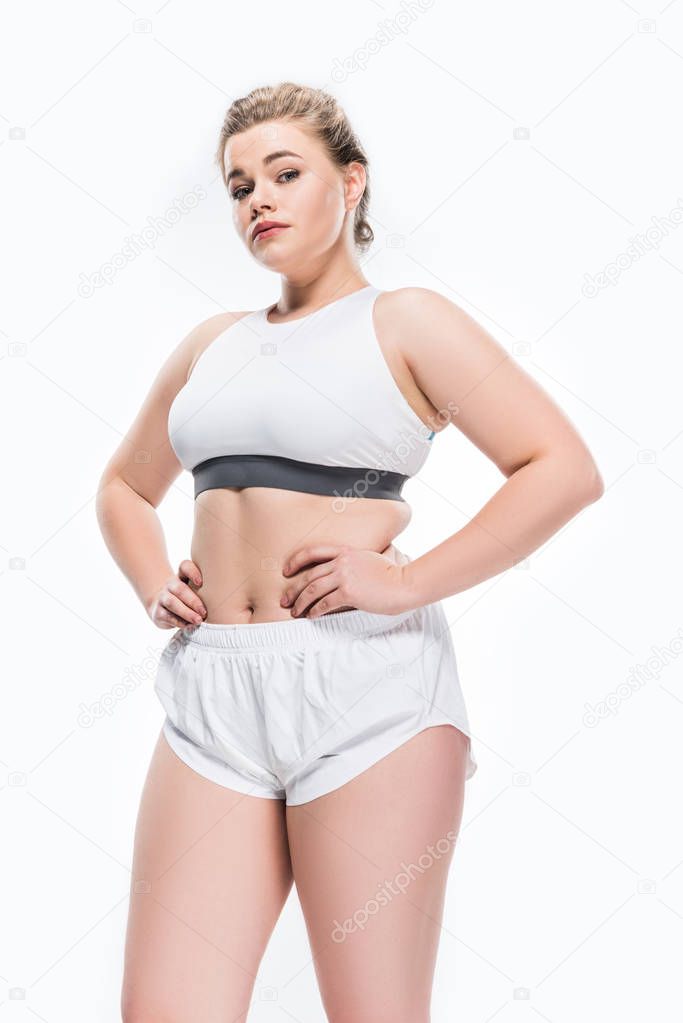low angle view of young overweight woman in sportswear standing with hands on waist and looking at camera isolated on white 