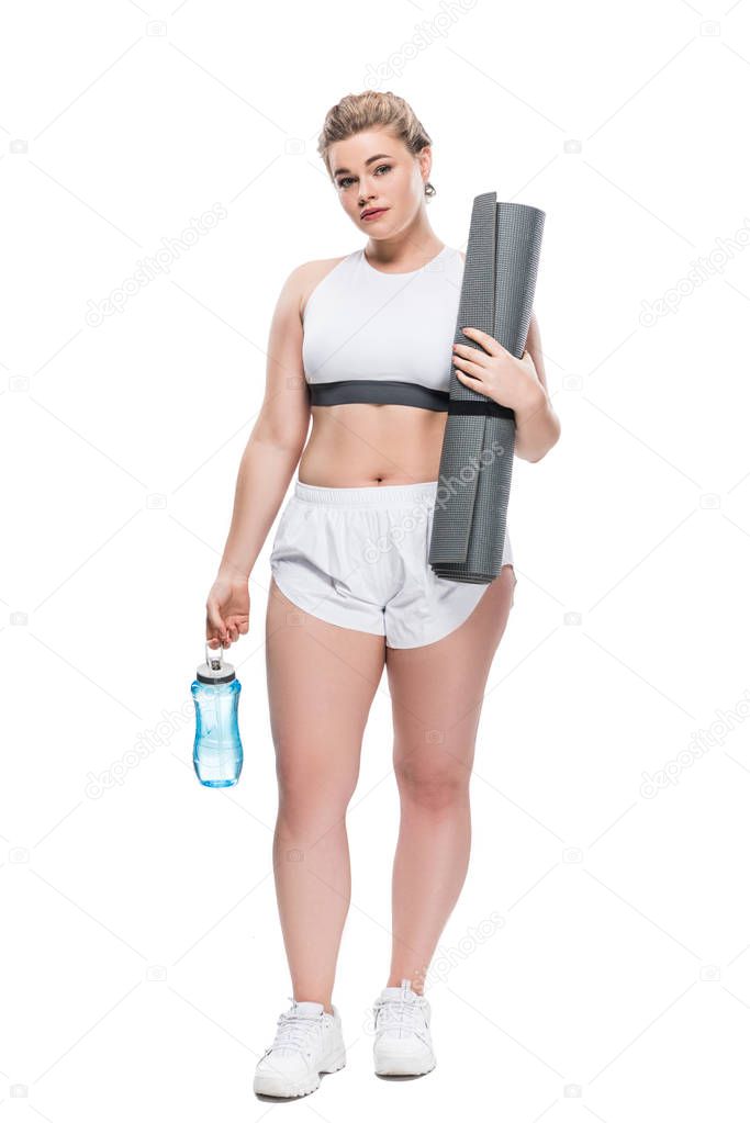 attractive oversize girl in sportswear holding bottle of water and yoga mat isolated on white