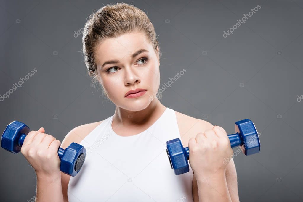 young woman training with dumbbells and looking away isolated on grey