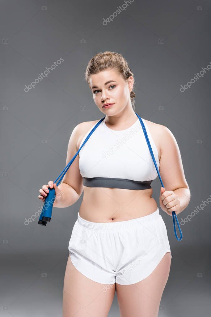 young oversize woman in sportswear holding skipping rope and looking at camera on grey