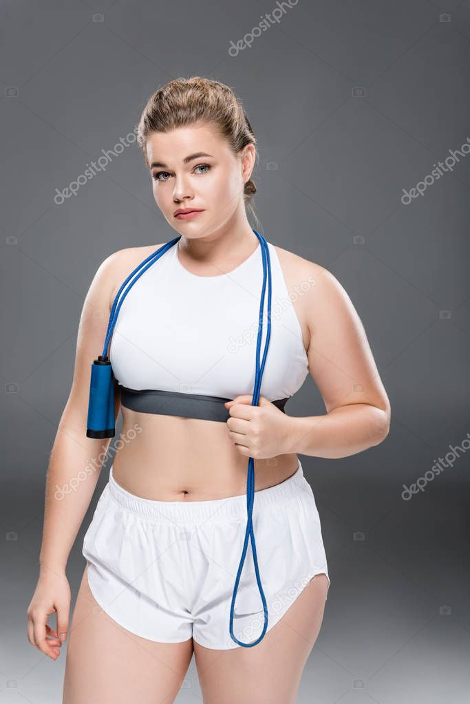 attractive young oversize woman in sportswear holding skipping rope and looking at camera on grey