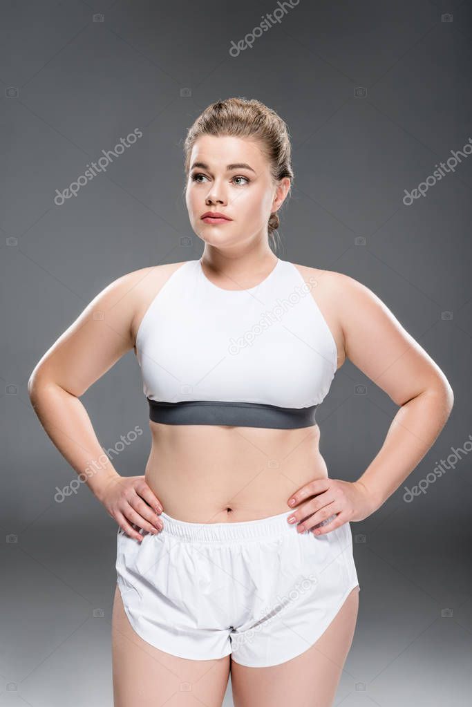 attractive young oversize woman in sportswear standing with hands on waist and looking away on grey