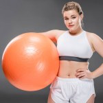 Mladá žena s nadváhou v sportovní hospodářství, fit ball a při pohledu na fotoaparát izolované Grey