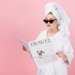 Young oversize woman in bathrobe, sunglasses and towel on head reading travel newspaper isolated on pink