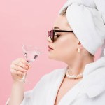 Jeune femme oversize en peignoir, lunettes de soleil et serviette sur la tête boire du verre isolé sur rose