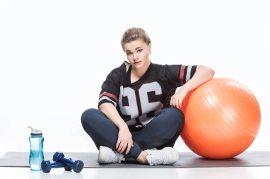 young overweight woman in sportswear leaning at fit ball and looking at camera while sitting on yoga mat isolated on white clipart