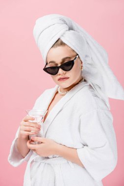 young oversize woman in bathrobe, sunglasses and towel on head holding glass isolated on pink clipart