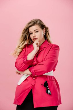 attractive size plus woman in fashionable jacket posing with hand on chin and looking at camera isolated on pink