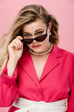 portrait of beautiful oversize girl adjusting sunglasses and looking at camera isolated on pink clipart