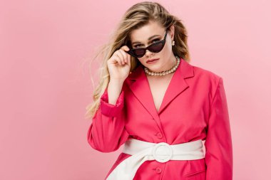 beautiful plus size woman in pink trench coat adjusting sunglasses and looking at camera isolated on pink clipart