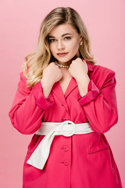 elegant young woman in pink trench coat looking at camera isolated on pink 