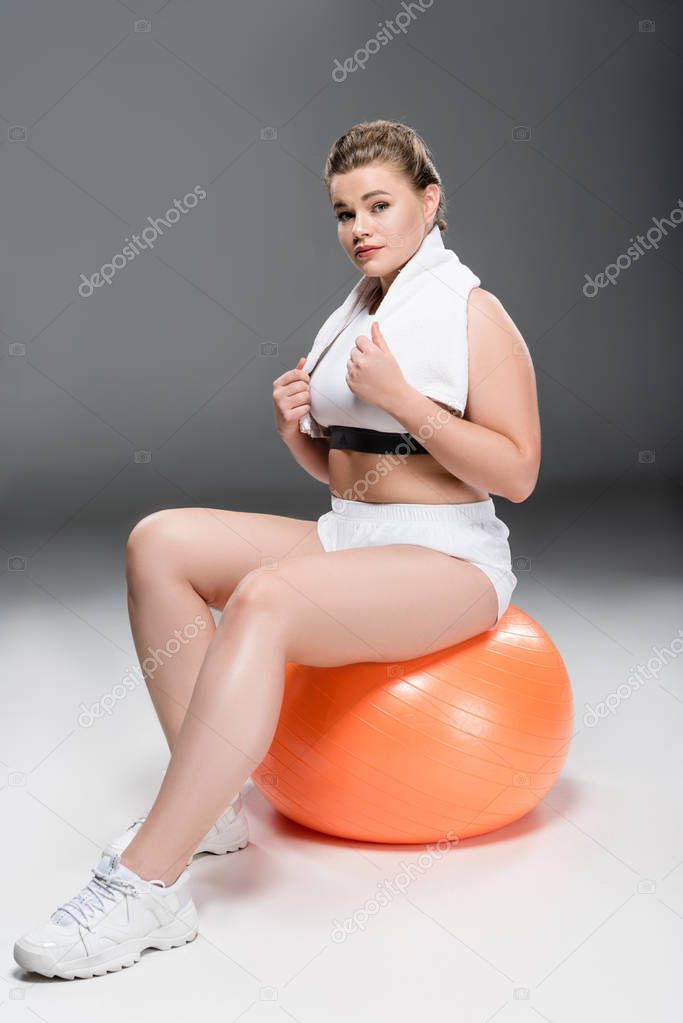 young oversize woman with towel sitting on fit ball and looking at camera on grey 