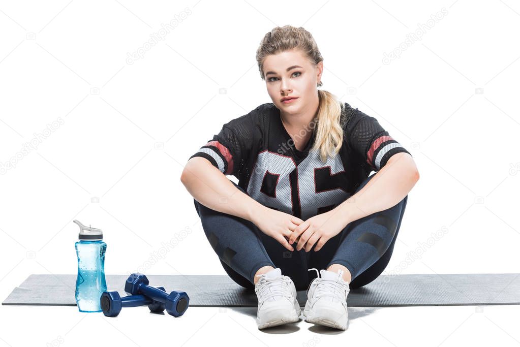 young size plus woman in sportswear sitting on yoga mat and looking at camera isolated on white  