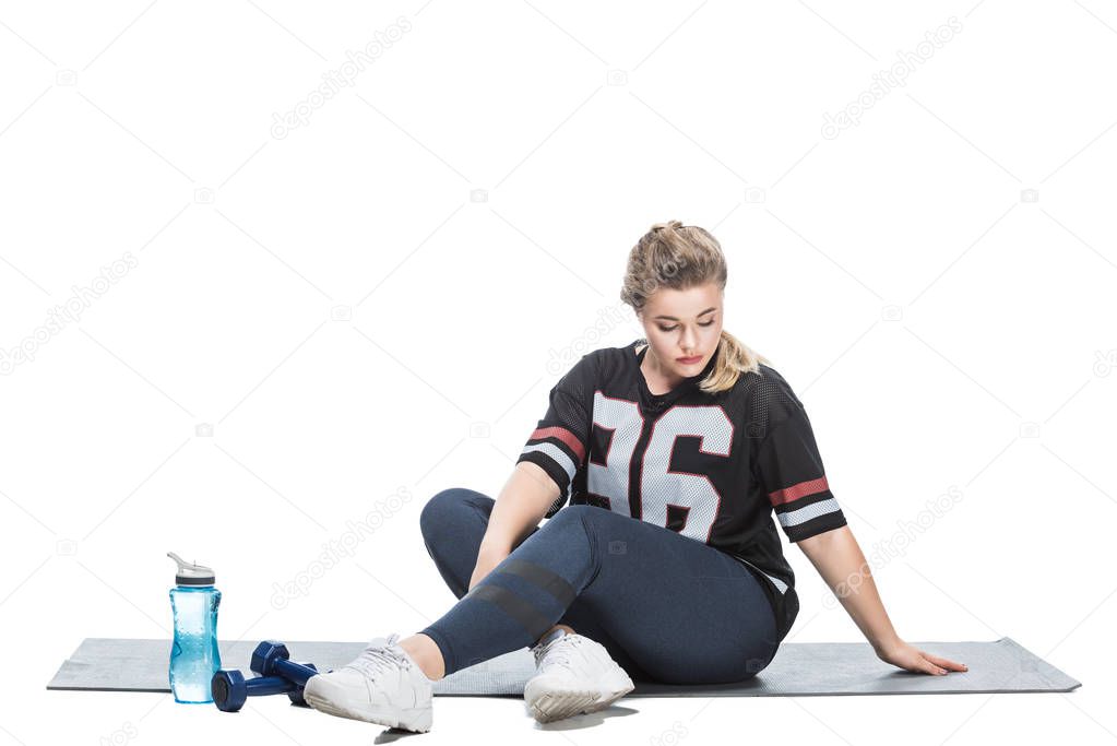 young overweight woman in sportswear sitting on yoga mat and looking down isolated on white 