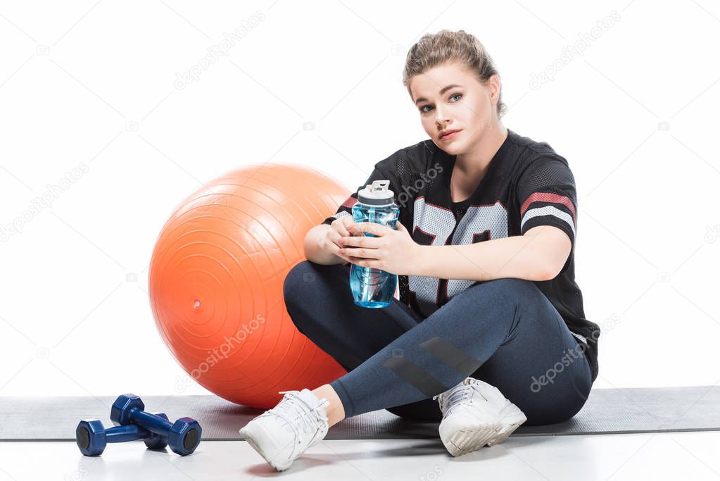 young overweight woman in sportswear holding bottle of water and looking at camera while sitting on yoga mat isolated on white