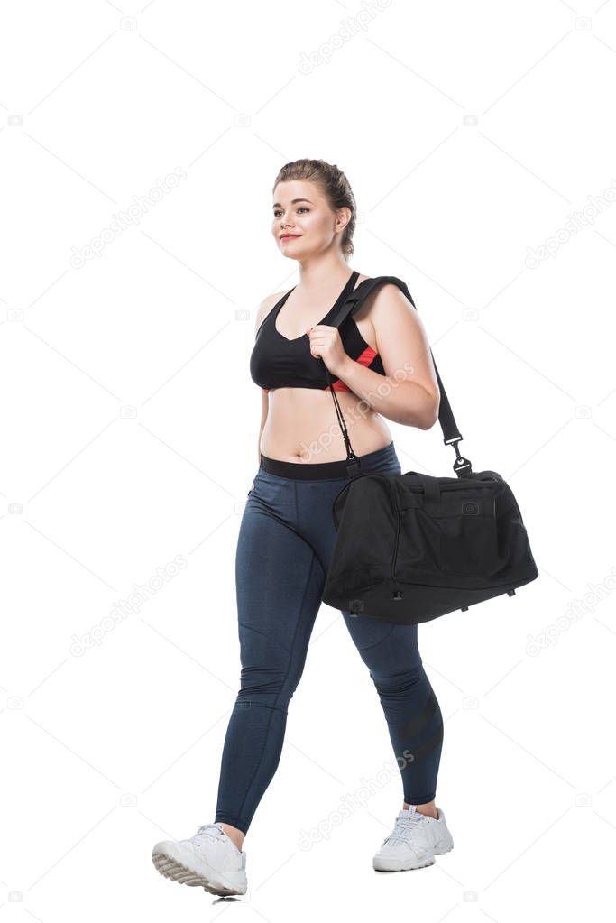 smiling young size plus woman with sports bag walking and looking away isolated on white