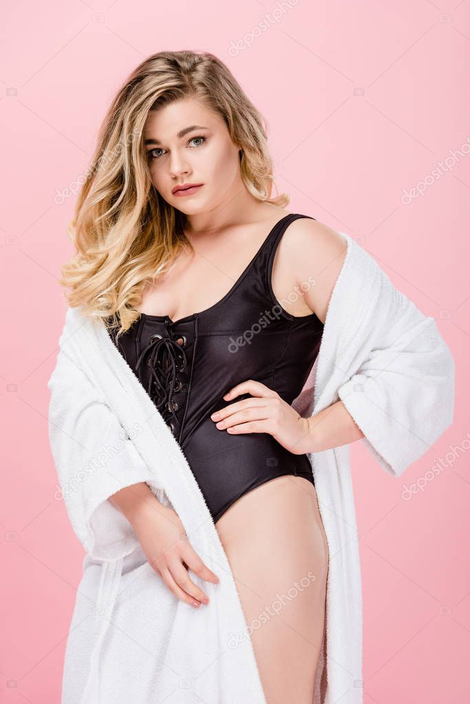 attractive overweight girl in swimsuit and bathrobe standing with hand on waist and looking at camera isolated on pink