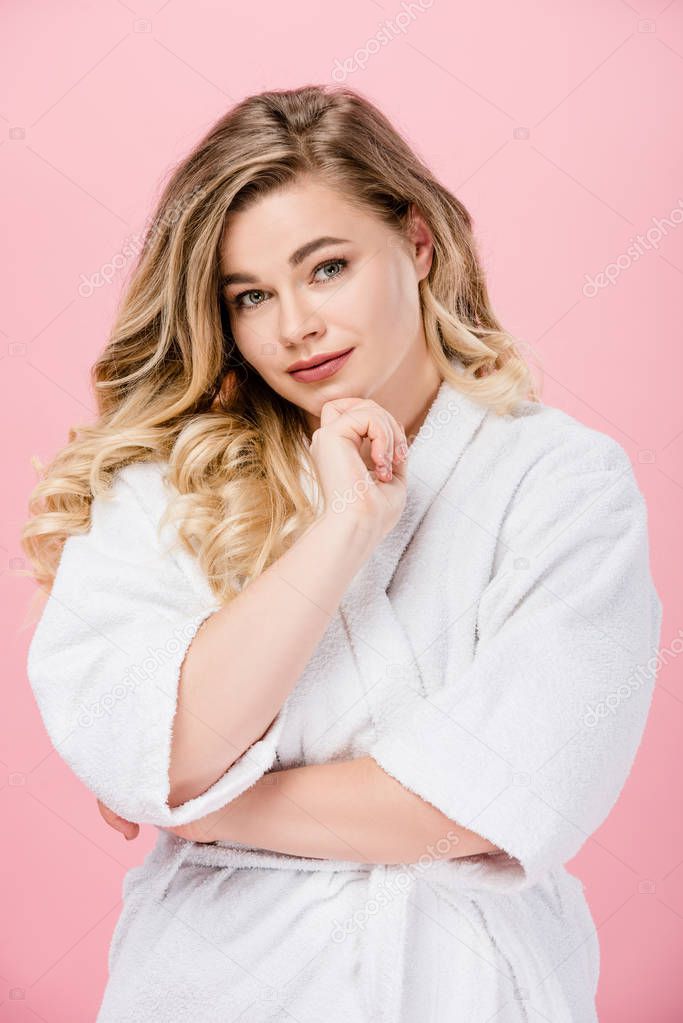 beautiful overweight girl in bathrobe standing with hand on chin and smiling at camera isolated on pink