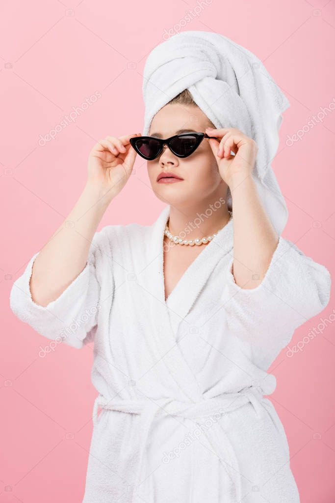 beautiful oversize girl in bathrobe and towel on head adjusting sunglasses isolated on pink