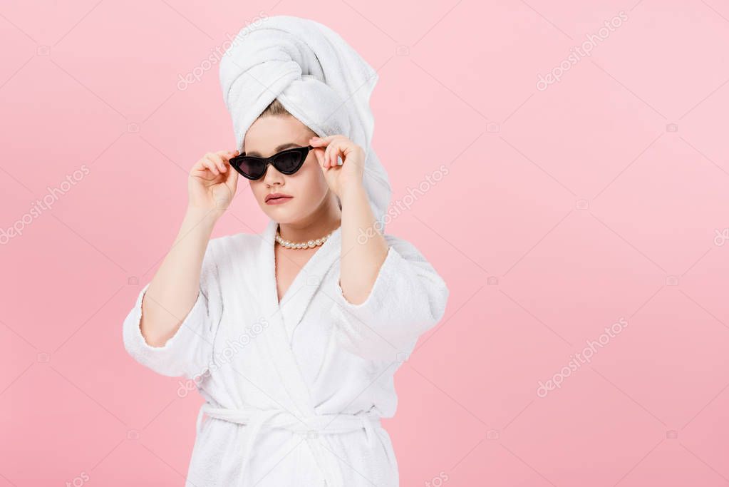 young oversize woman in bathrobe and towel on head adjusting sunglasses isolated on pink
