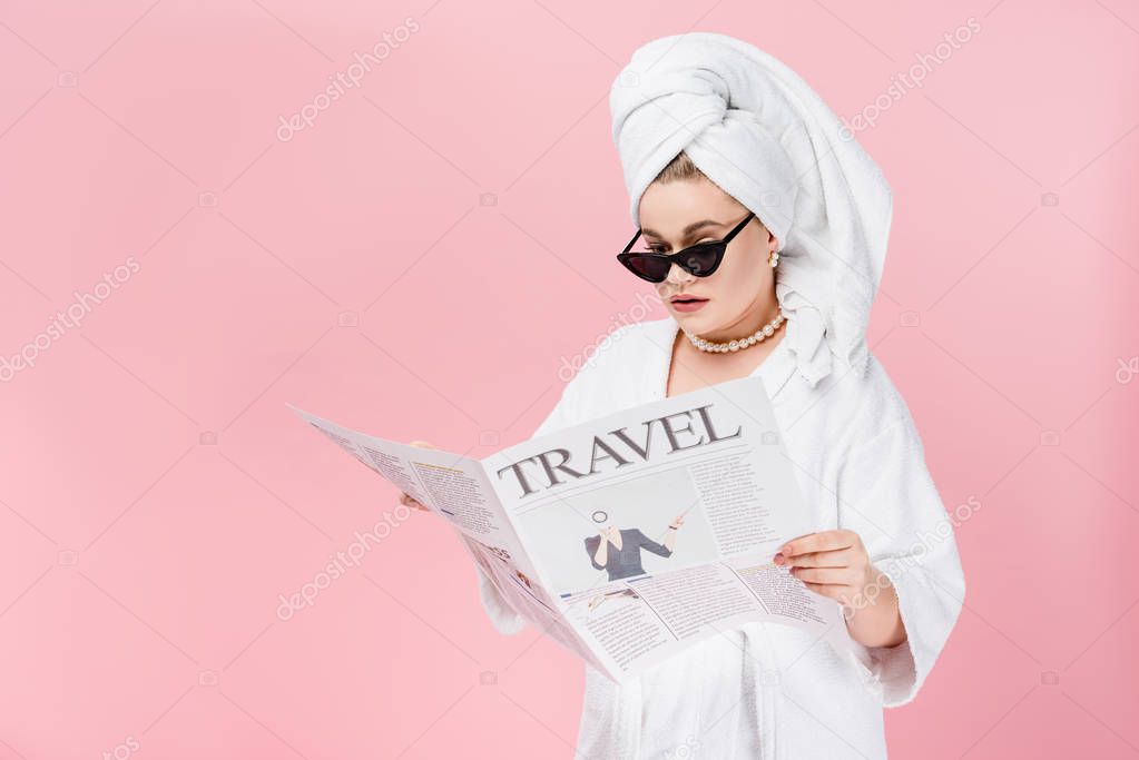young size plus woman in bathrobe, sunglasses and towel on head reading travel newspaper isolated on pink