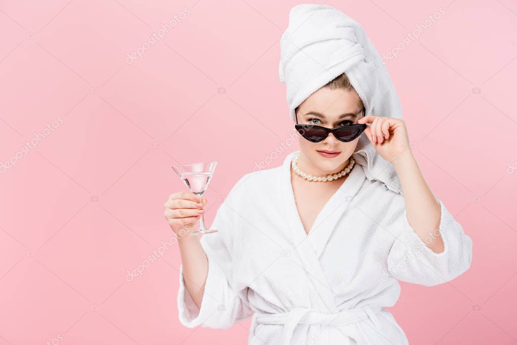 attractive oversize woman in bathrobe, sunglasses and towel on head holding glass and looking at camera isolated on pink