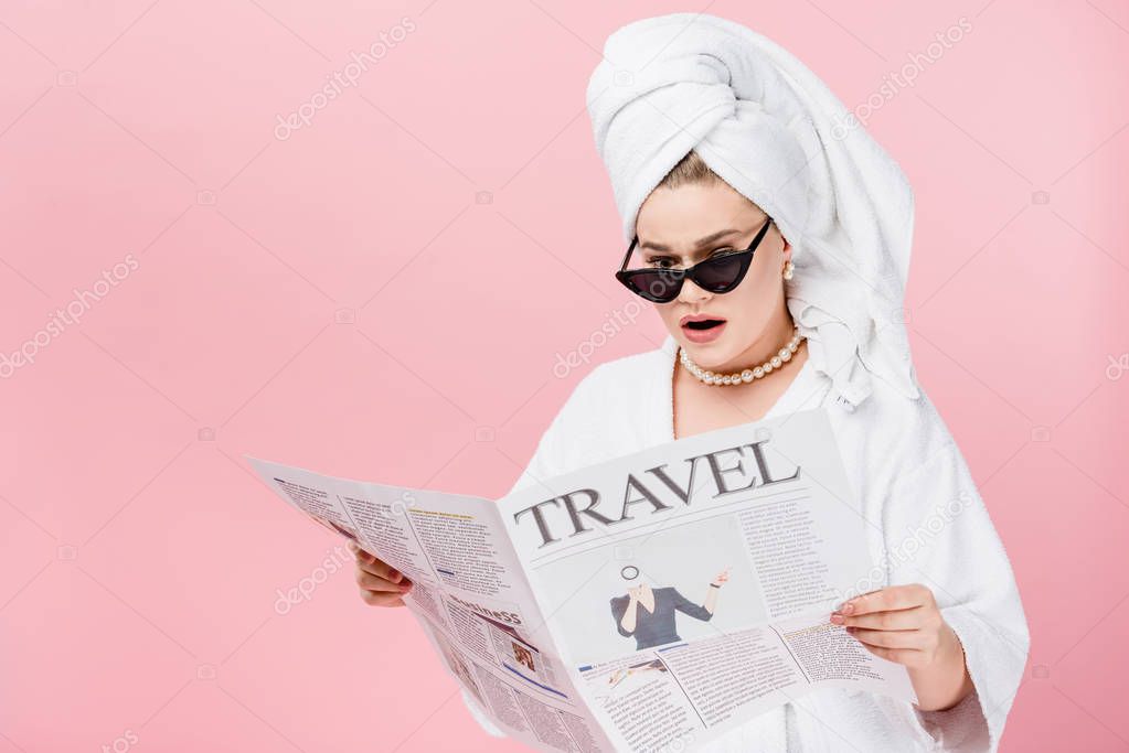 shocked young size plus woman in bathrobe, sunglasses and towel on head reading travel newspaper isolated on pink