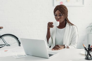 Afrika kökenli Amerikalı yetişkin iş kadını bilgisayar masa başında oturup, kahve holding ve ofiste çalışan beyaz takım