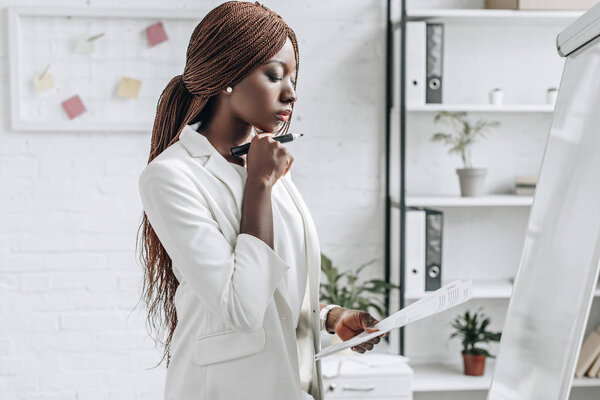 pensive african american adult businesswoman in white formal wear holding document and working on project near flip chart in office