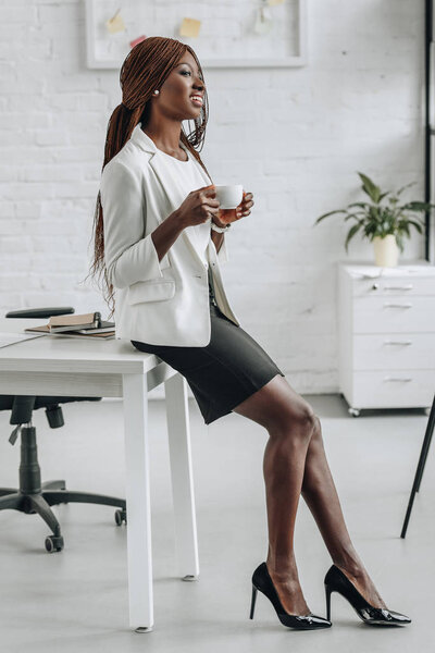 african american adult businesswoman in white formal wear leaning on office desk, smiling and holding cup of coffee