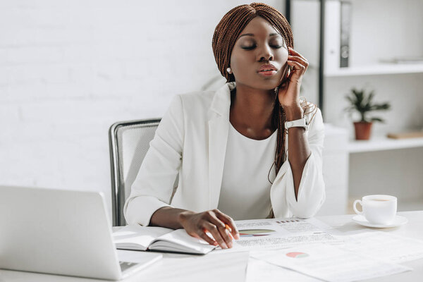 tired african american adult businesswoman with closed eyes working on project and sitting at computer desk with cup of coffee
